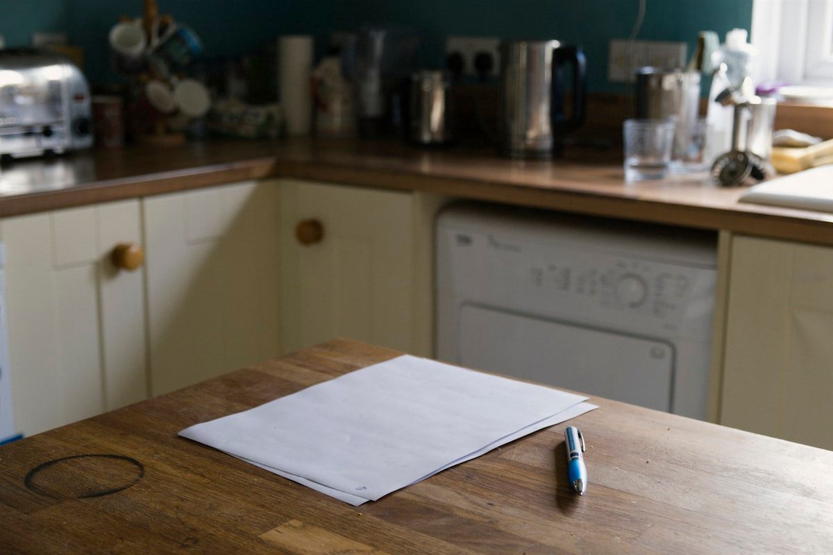 Two moving estimate sheets laid side by side on a kitchen table, one with a clipped, boxed total and the other with several penciled-in add-on charges, alongside a stack of household inventory notes and a calculator.