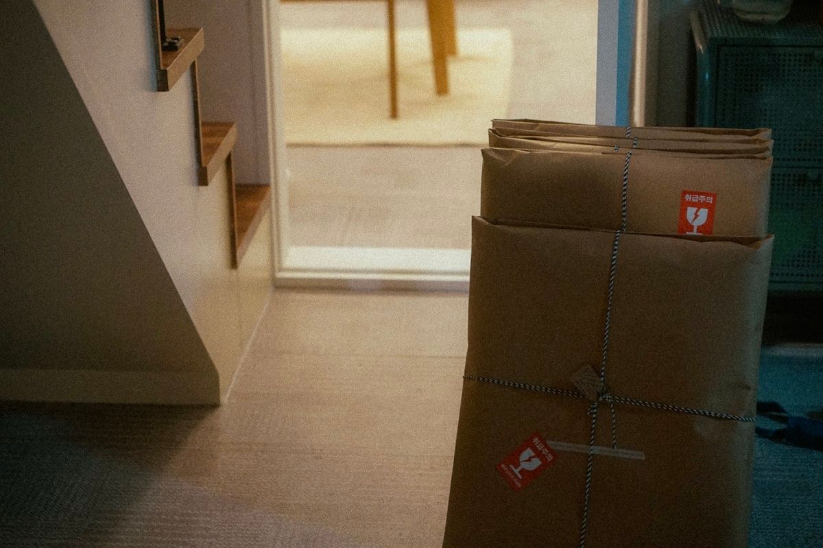 A packed living room with moving boxes stacked near a couch, rolls of packing tape, labeled markers, a tape gun, and a coffee maker still plugged into a wall outlet, showing a move in progress