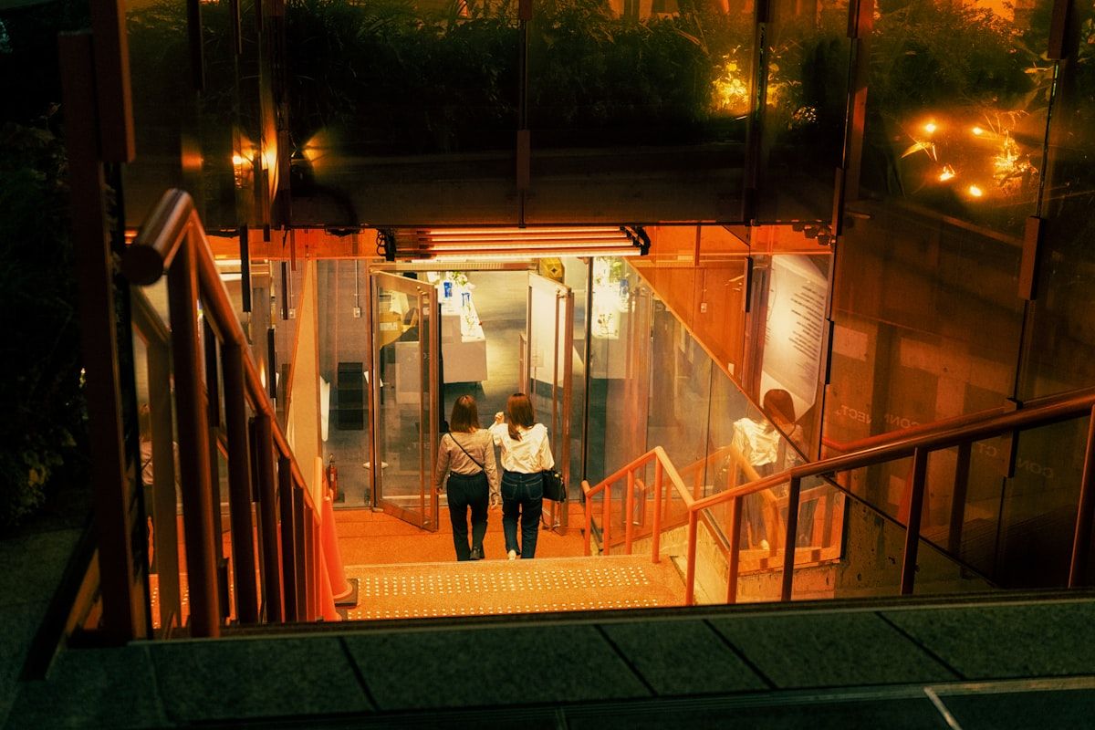 A narrow condo stairwell with movers carrying a sofa upward, a service elevator waiting at the landing, and a small shuttle truck parked farther down the street, suggesting extra access-related moving charges.