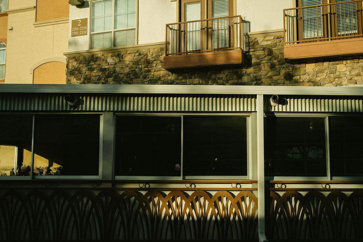 A moving truck parked beside a condo building with an elevator entrance and stacked moving blankets, hand trucks, taped boxes, and a couple of movers carrying a sofa through a tight hallway while a frustrated resident watches from the side