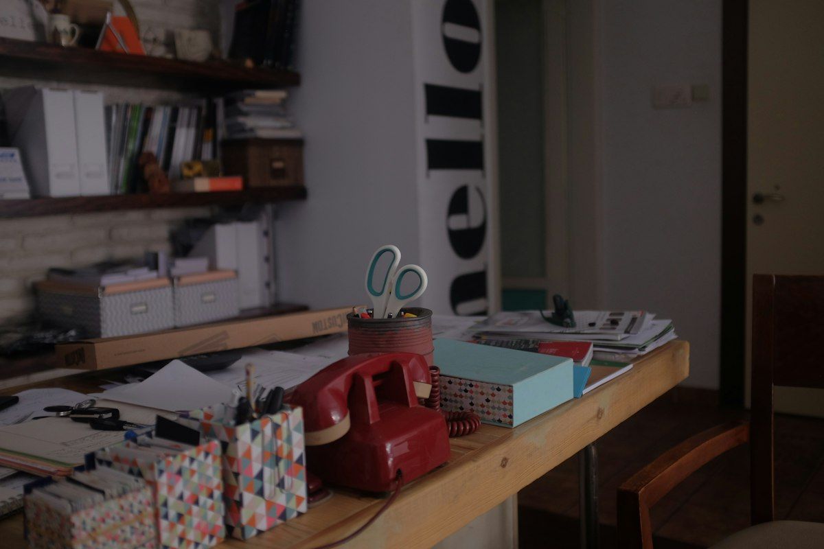 A kitchen table covered with three printed moving estimates, a clipboard, a tape measure, a small box of packing materials, and a person comparing the papers while looking at a partially packed room with labeled boxes and a wrapped lamp